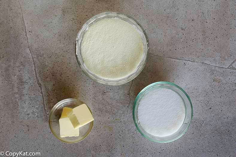 An overhead view of ingredients for sweetened condensed milk in glass bowls: powdered milk, sugar, and two pats of butter.