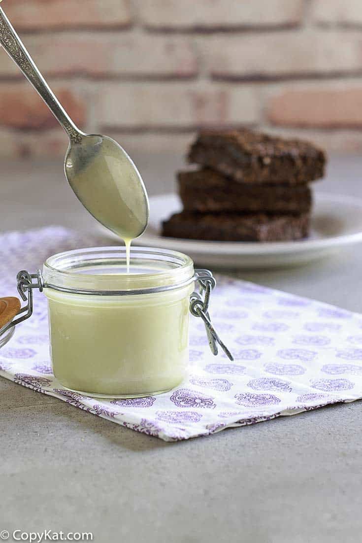 A spoon drips homemade sweetened condensed milk into a glass jar. A plate of brownies sits out of focus in the background.