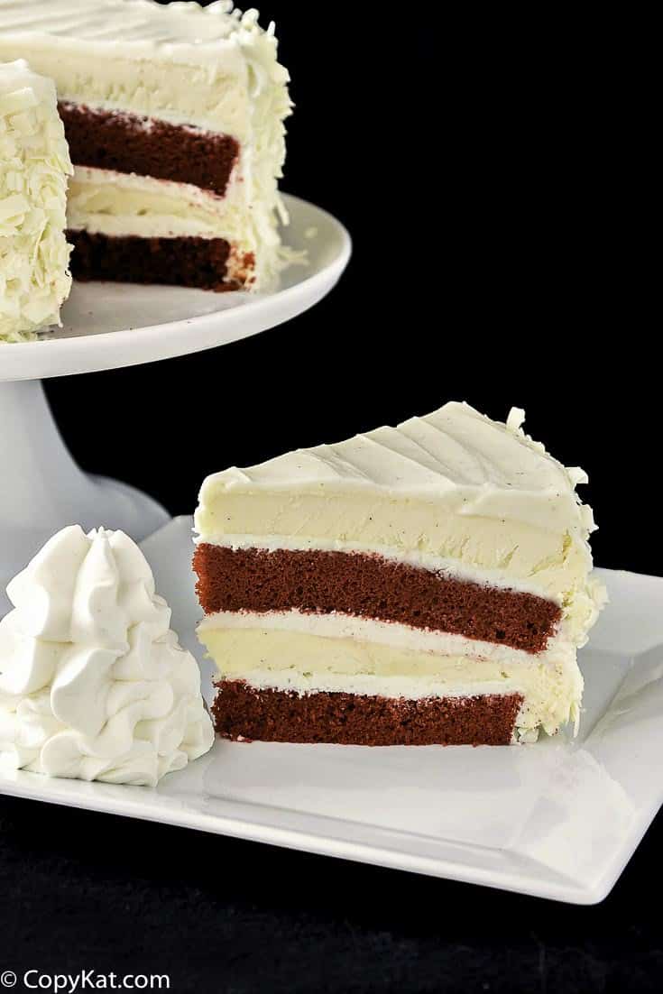 A slice of homemade Cheesecake Factory Red Velvet Cheesecake on a plate next to the cake on a stand