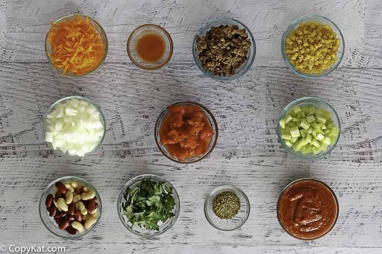 An overhead, flat-lay view of the many ingredients for Olive Garden's Pasta e Fagioli soup. The ingredients, including cooked ground beef, pasta, beans, and chopped vegetables, are neatly arranged in individual glass bowls.