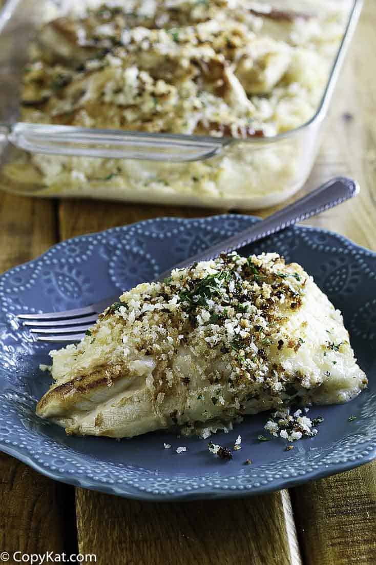 A plated piece of copycat Longhorn parmesan crusted chicken on an embossed blue plate. The golden-brown panko and herb topping is in sharp focus, with the full baking dish visible in the background.