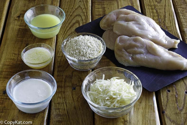 n overhead view of the ingredients for parmesan crusted chicken on a wooden table. Pictured are several raw chicken breasts and glass bowls containing panko breadcrumbs, shredded cheese, grated parmesan, cream, and a liquid base.