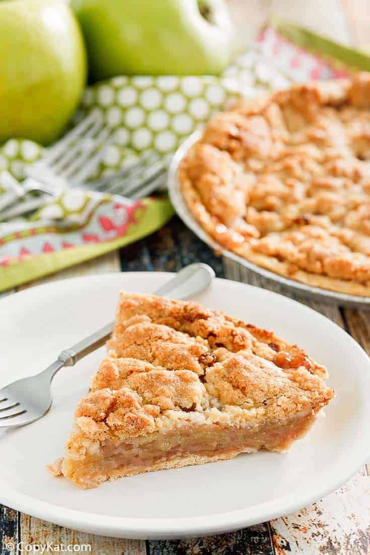 a slice of Dutch apple pie and a fork on a plate with the pie and apples in the background