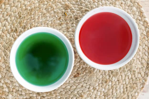 Two white bowls containing liquid red and green Jello for filling the cake.