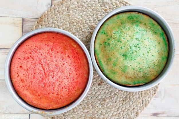 Two round white cake layers soaked in red and green liquid Jello in baking pans.