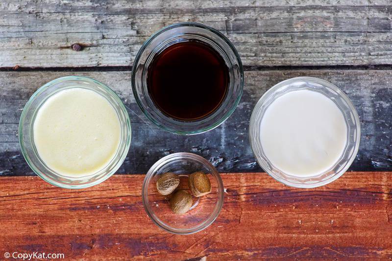 An overhead view of the ingredients for a Starbucks eggnog latte on a rustic wood background. Pictured are four glass bowls containing espresso, milk, eggnog, and whole nutmegs.