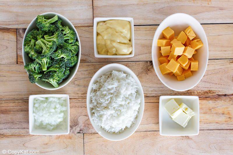 An overhead shot of the ingredients for broccoli cheese rice casserole arranged in white bowls on a wooden surface. Pictured are bowls of fresh broccoli florets, cooked white rice, diced onion, cubed Velveeta cheese, cream of chicken soup, and butter.