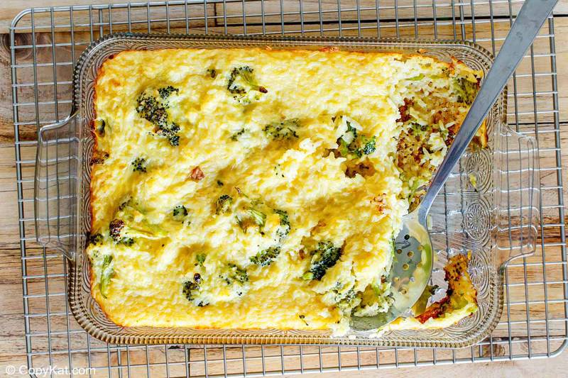 An angled, top-down view of the finished broccoli cheese rice casserole in a glass baking dish. The casserole is golden and bubbly, and a serving has been removed with a metal spoon, showcasing the cheesy rice and broccoli inside.