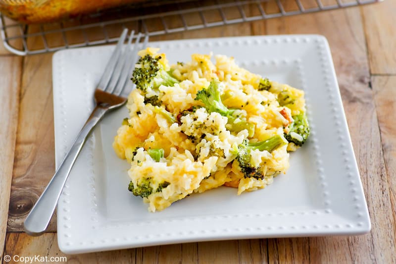 A close-up of a serving of broccoli cheese rice casserole on a square white plate. The portion clearly shows the creamy blend of rice, cheese, and bright green broccoli florets, with a fork resting beside it.