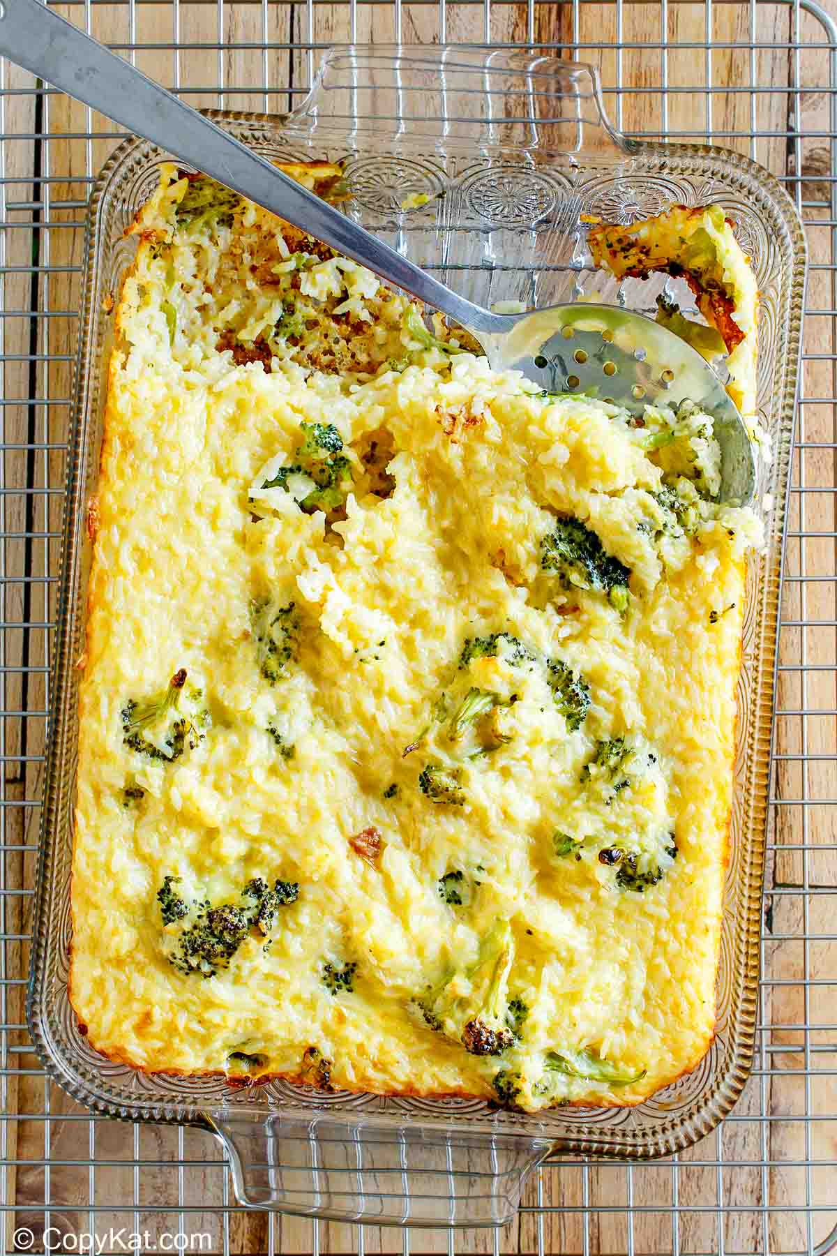 An angled, top-down view of the finished broccoli cheese rice casserole in a glass baking dish. The casserole is golden and bubbly, and a serving has been removed with a metal spoon, showcasing the cheesy rice and broccoli inside.