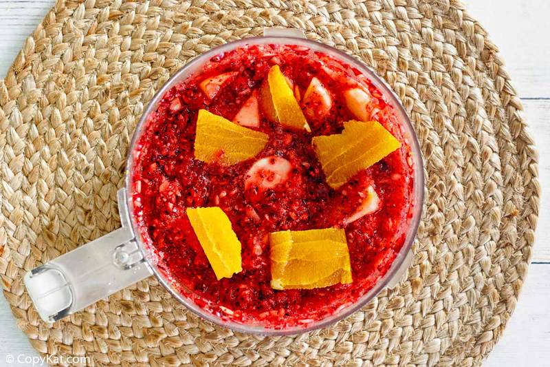 A top-down view into a food processor showing the ingredients for cranberry relish being pureed. The bright red, textured mixture has chunks of fresh orange on top, ready for the final processing.
