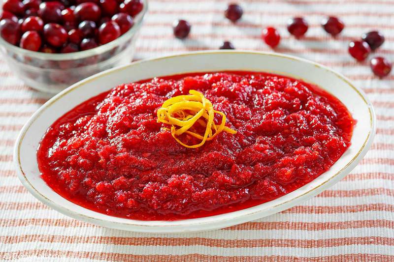 A white oval dish filled with vibrant, fresh cranberry relish, garnished with a swirl of orange zest. A bowl of whole cranberries is visible in the background, highlighting the main ingredient.