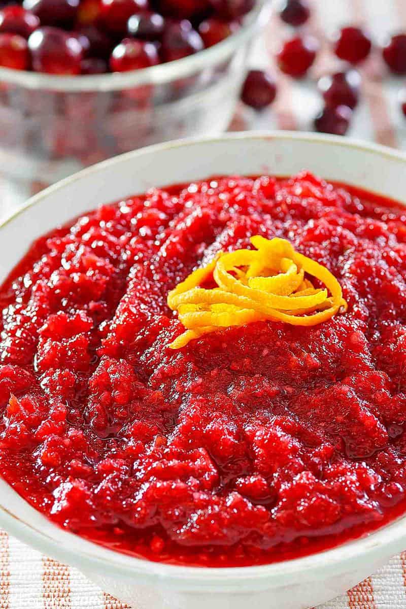 A close-up of a bowl of fresh cranberry relish, focusing on its vibrant red color and coarse texture. The relish is topped with a beautiful garnish of orange zest.