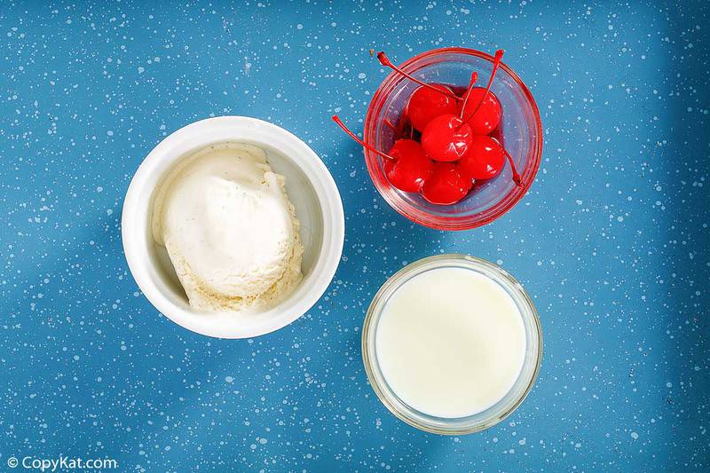 Overhead view of eggnog shake ingredients: vanilla ice cream, eggnog, and maraschino cherries in glass bowls on a blue surface.
