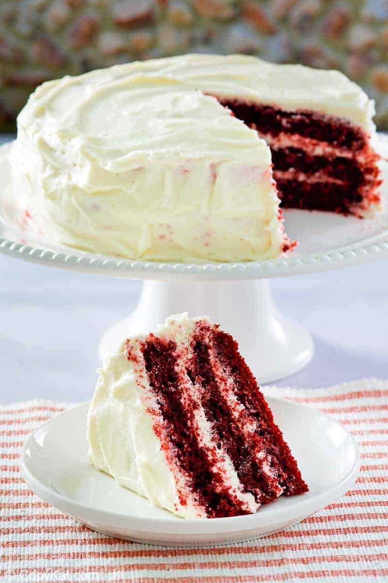 homemade Waldorf Astoria Red Velvet Cake slice on a plate in front of the cake on a stand