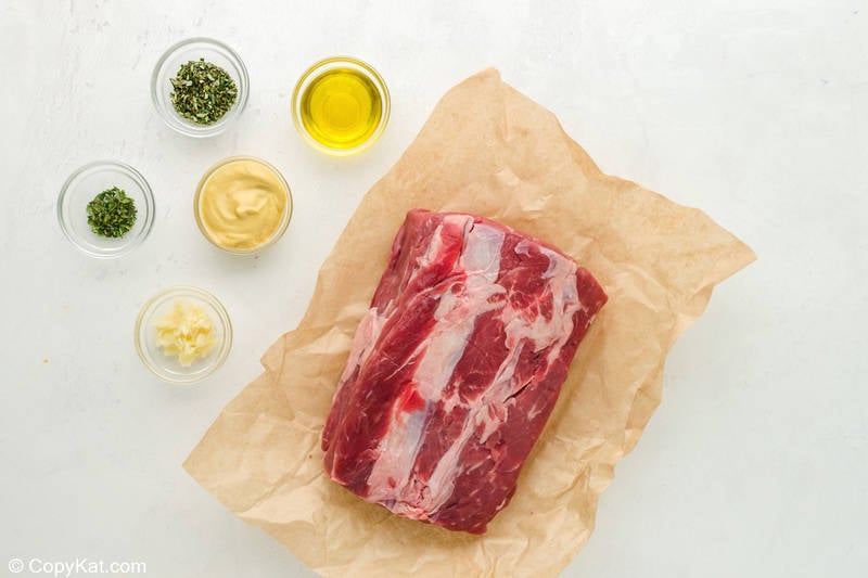 An overhead view of the ingredients for a New York Strip roast. Pictured are a large, raw strip loin roast on butcher paper and small glass bowls containing olive oil, Dijon mustard, minced garlic, fresh rosemary, and fresh thyme.