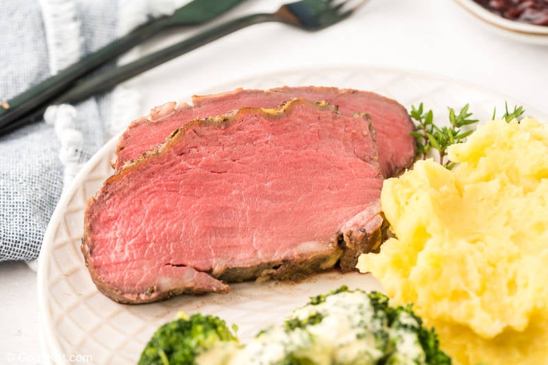 A plated meal featuring two thick slices of tender, pink New York Strip roast served with a side of creamy mashed potatoes and broccoli with cheese sauce.