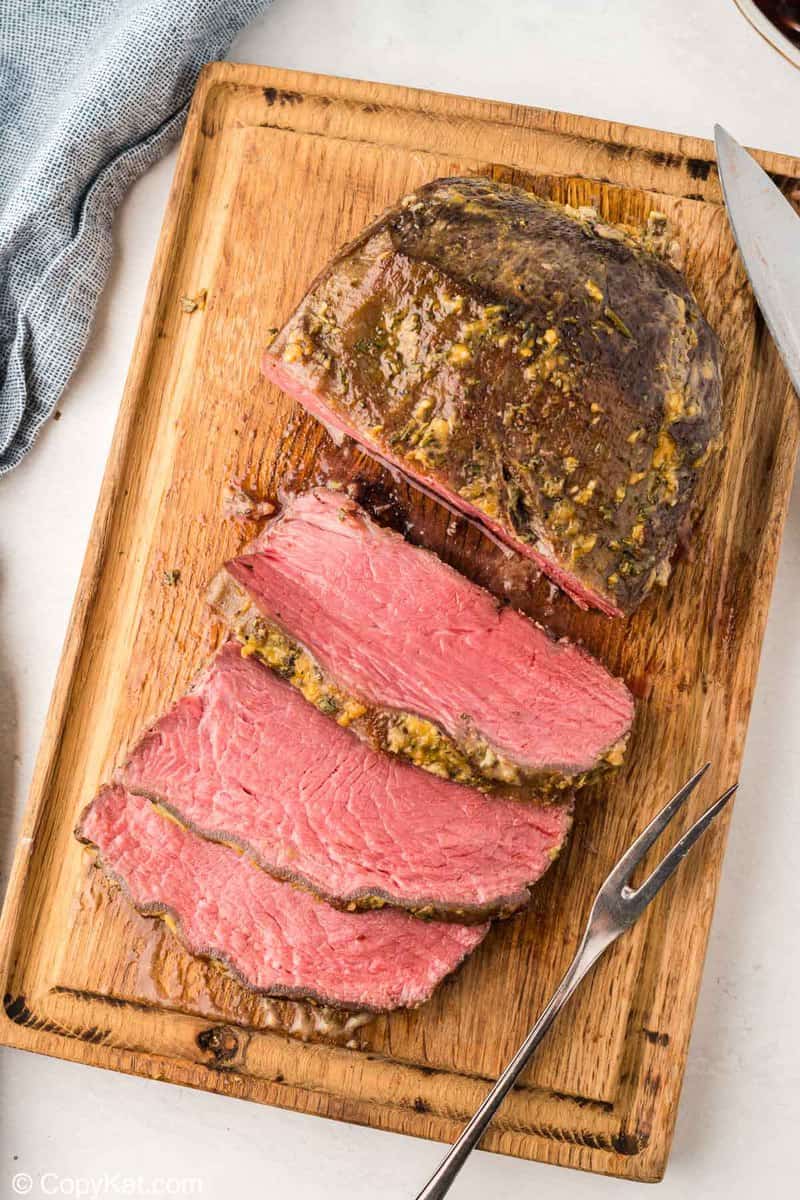 A cooked New York Strip roast with a dark herb crust being carved on a wooden cutting board. Several thick slices have been cut, revealing the juicy, pink, medium-rare center of the roast.