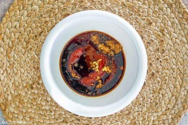 An overhead shot of a white bowl containing the unmixed ingredients for tonkatsu sauce. A dark liquid base is topped with ketchup, brown sugar, minced ginger, and garlic, ready to be stirred.