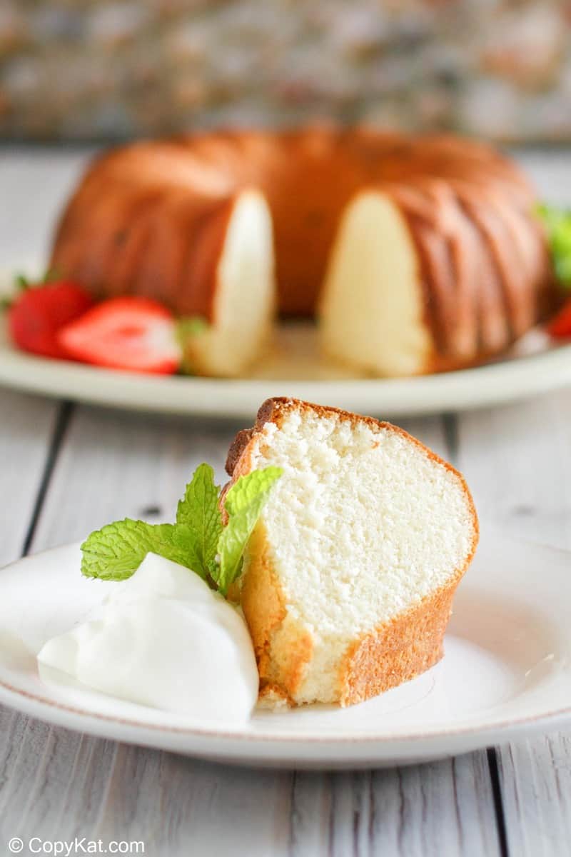 A close-up of a slice of Scandinavian almond cake on a plate, garnished with whipped cream and mint, with the bundt cake behind.