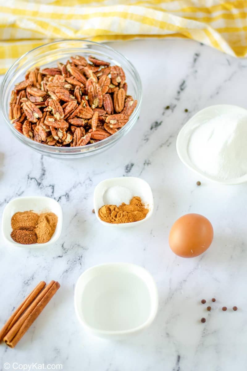 An overhead view of the ingredients for spiced pecans. Pictured are a large bowl of pecan halves, a whole egg, and smaller bowls containing water, sugar, and a variety of ground spices like cinnamon and nutmeg.