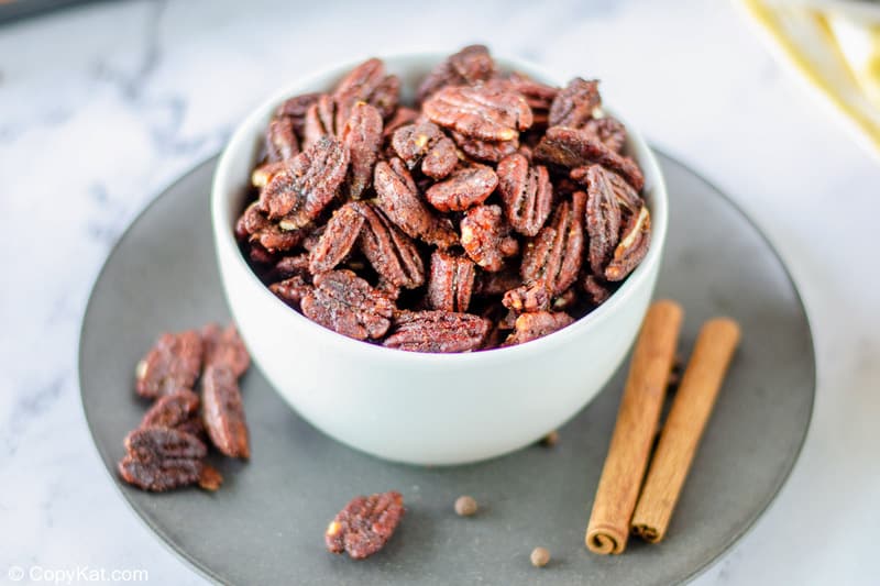 A white serving bowl filled with crispy, roasted spiced pecans. The bowl sits on a dark plate, garnished with cinnamon sticks, ready to be served as a snack.