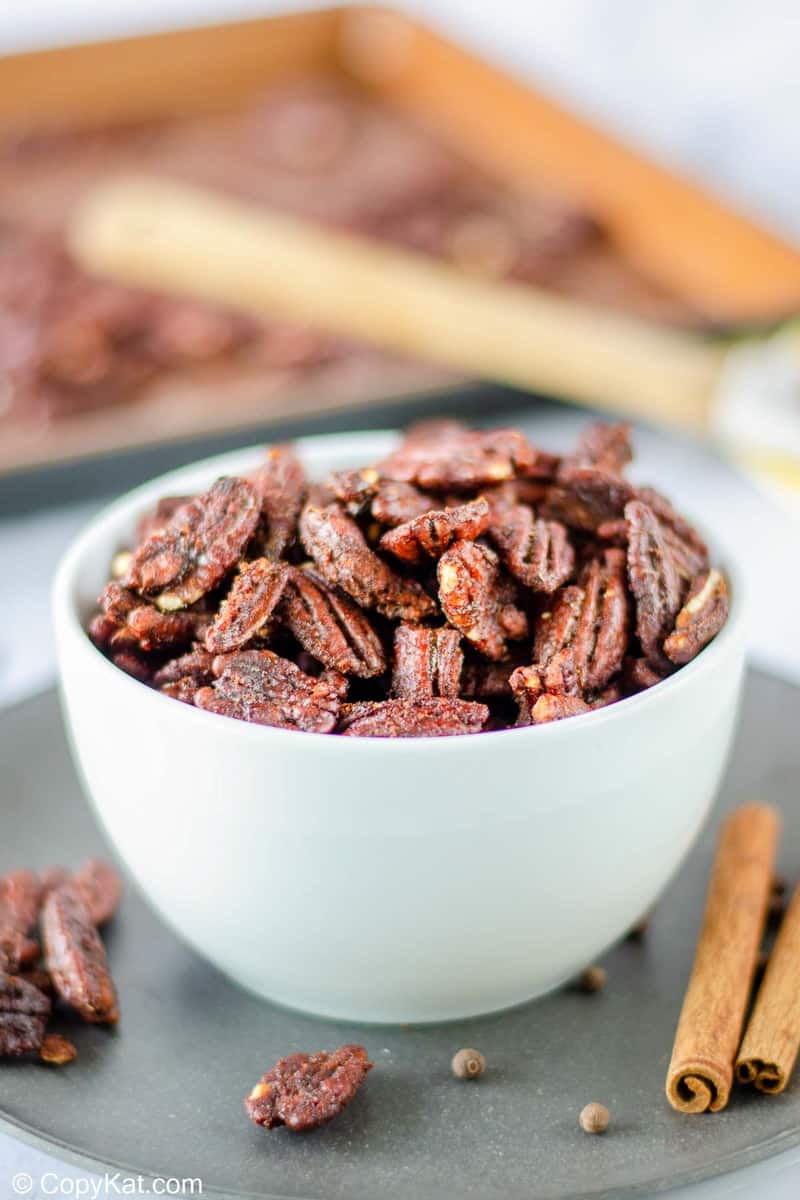 A close-up of a white bowl of spiced pecans, with the baking sheet used to roast them visible in the blurred background.