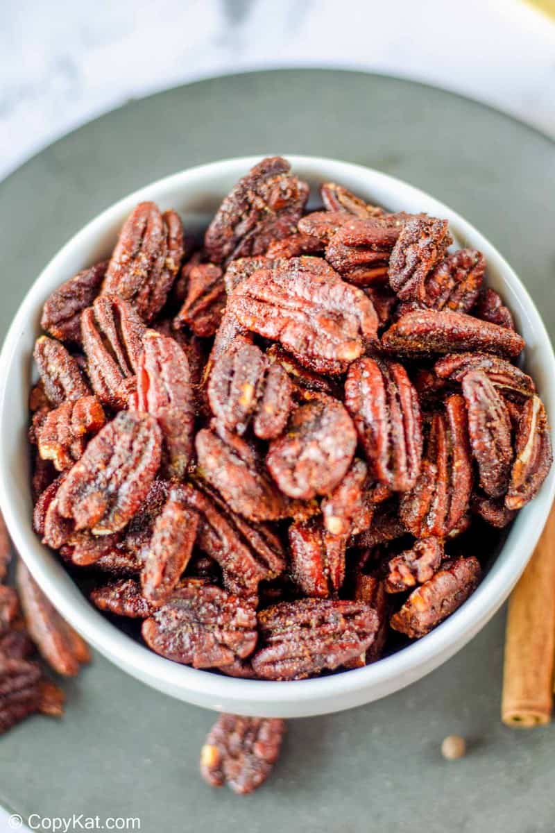 A close-up, overhead shot of a white bowl overflowing with freshly roasted spiced pecans, highlighting their deep color and sugary, spiced coating.