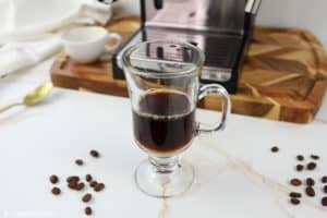 A clear glass mug containing two shots of dark, freshly brewed espresso, representing the first step in making a flat white. An espresso machine is visible in the background.