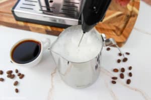 A close-up action shot of milk being frothed in a metal pitcher using a handheld frother. The milk has a thick, white microfoam on top, ready to be poured into the nearby cup of espresso.