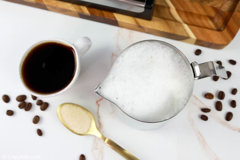 An overhead shot of the ingredients for a flat white on a marble counter. Pictured are a cup of espresso, a metal pitcher of frothed milk, and a spoonful of sugar, with an espresso machine in the background.