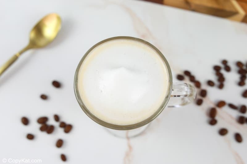 A direct overhead view of a finished flat white coffee, focusing on the smooth, perfect circle of white microfoam that tops the beverage in a clear glass mug.