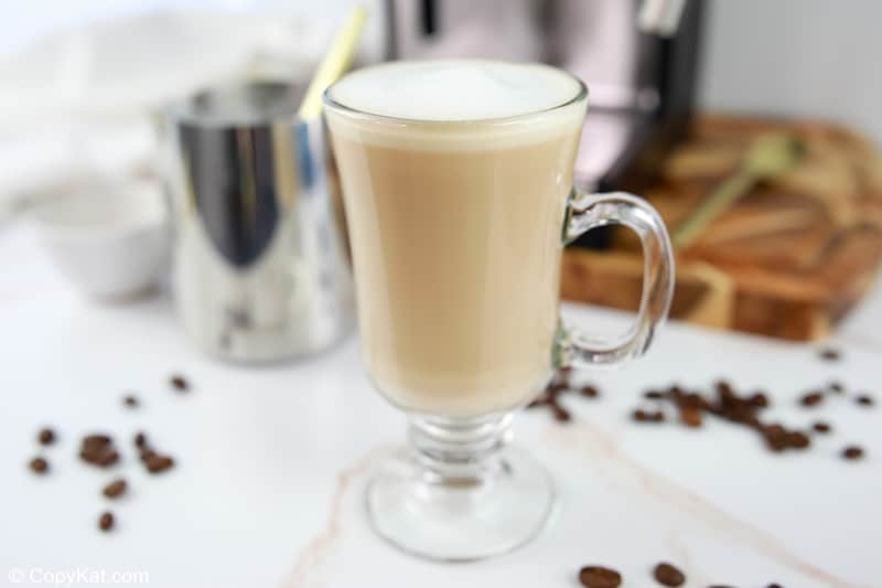 The finished Starbucks flat white served in a clear glass mug, showing its creamy, light-brown color and thin layer of foam. A milk pitcher and espresso machine sit in the background.