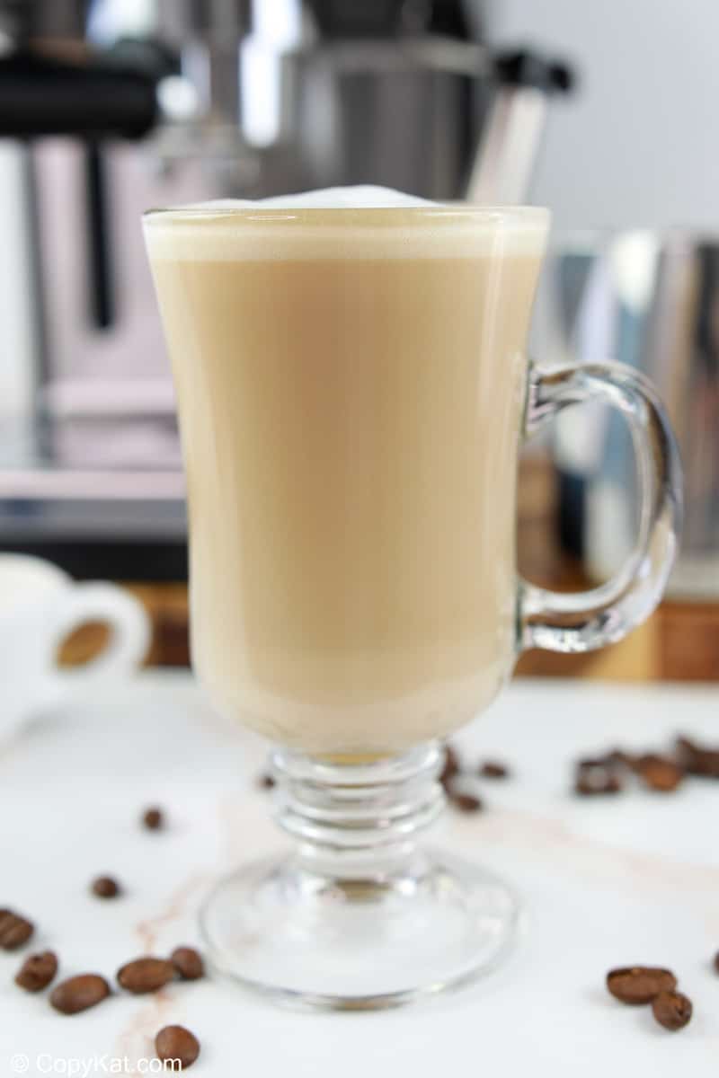 A homemade Starbucks flat white served in a tall, clear glass mug. The creamy coffee is topped with white foam, and an espresso machine is visible in the soft-focus background, creating an at-home barista scene.