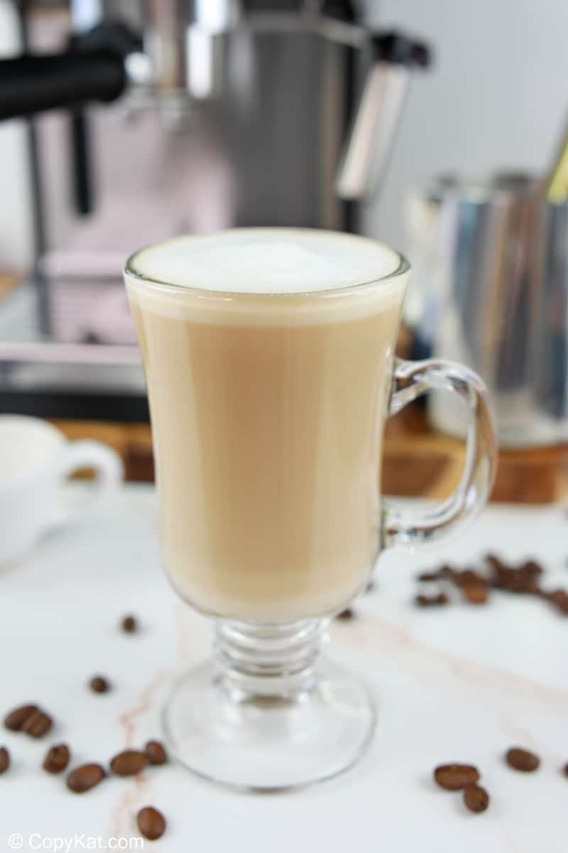 The finished Starbucks flat white served in a clear glass mug, showing its creamy, light-brown color and thin layer of foam. A milk pitcher and espresso machine sit in the background.