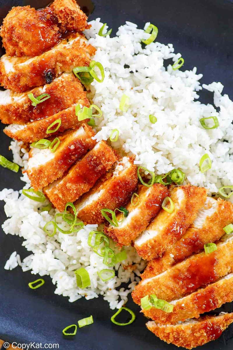A close-up, overhead view of sliced chicken katsu served on a bed of fluffy white rice. The image highlights the crispy texture of the panko crust, the dark drizzle of tonkatsu sauce, and the fresh green onion garnish.
