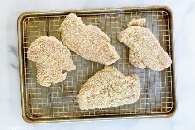Four pieces of chicken katsu, fully coated in panko breadcrumbs, are arranged on a wire rack, ready for frying.