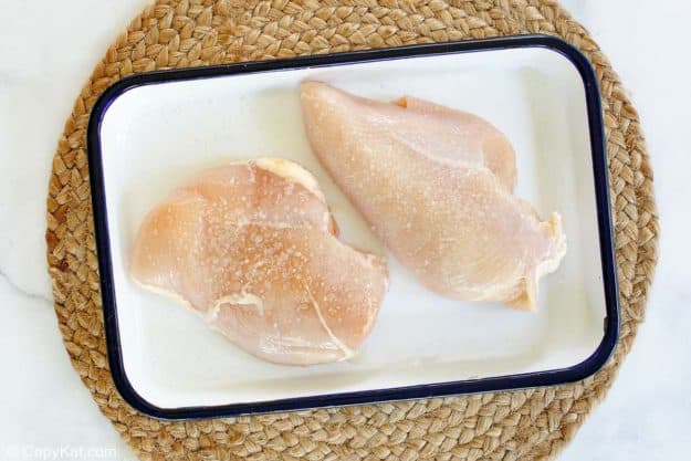 Two raw, thin chicken cutlets sprinkled with salt, resting in a white enamel dish as part of the preparation for making chicken katsu.