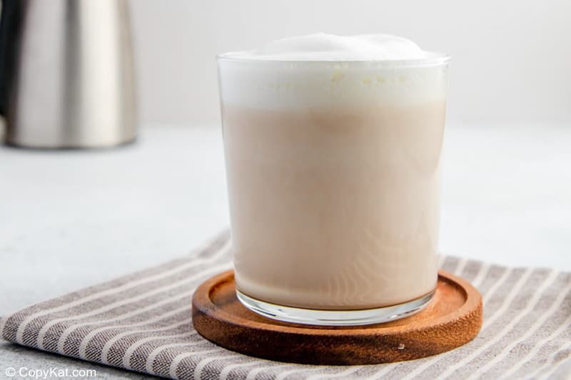 A homemade Starbucks London Fog served in a clear glass on a wooden coaster. The creamy tea is topped with a thick layer of white milk foam, with a frothing pitcher visible in the background.