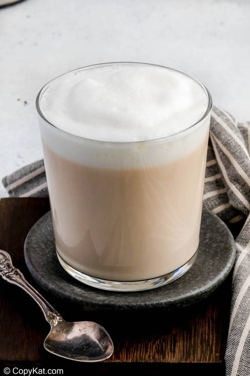 A finished London Fog tea latte in a clear glass, topped with a generous layer of milk foam. The drink is elegantly styled on a dark coaster with an antique spoon and a napkin.