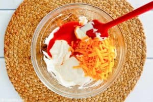 An overhead view of a glass mixing bowl containing softened cream cheese, shredded cheddar, and Frank's RedHot sauce. A red spatula is poised to stir the ingredients for the buffalo chicken dip together.