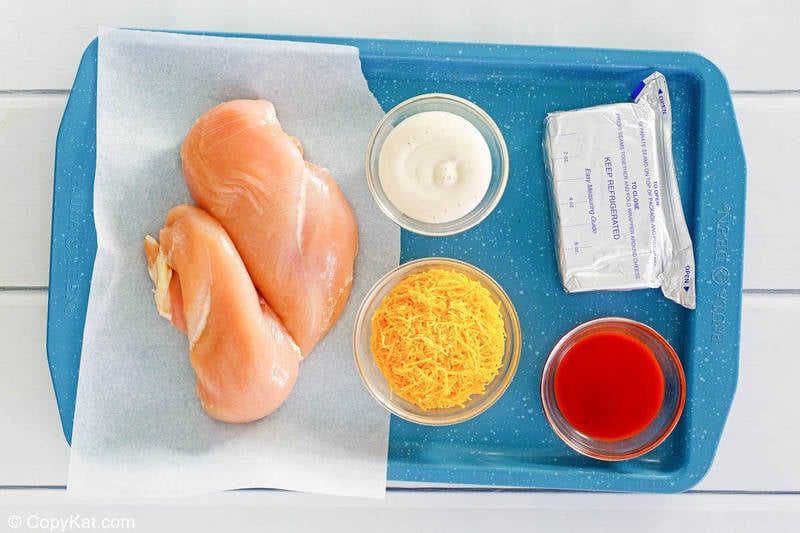 An overhead view of the five ingredients for Frank's buffalo chicken dip arranged on a blue tray. Pictured are two raw chicken breasts, a block of cream cheese, and bowls of shredded cheddar cheese, ranch dressing, and Frank's RedHot Sauce.