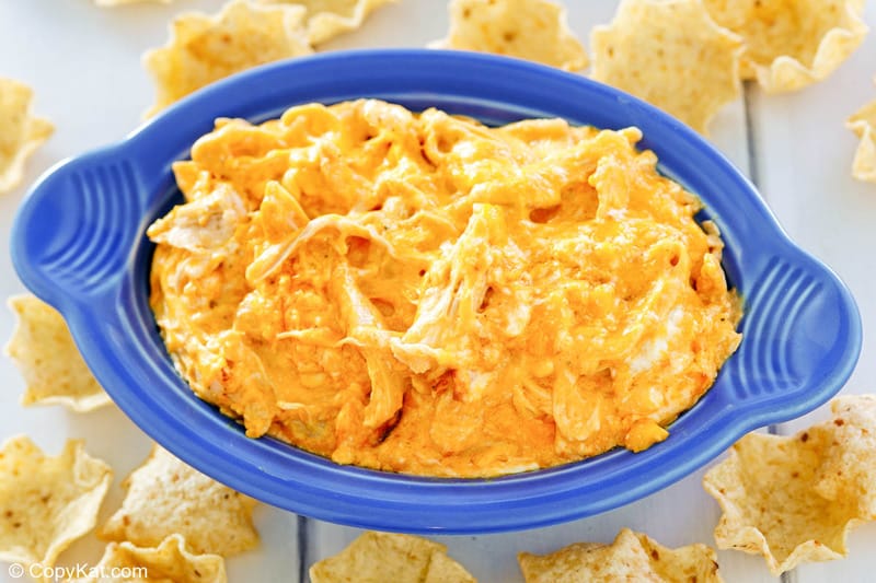 An angled, close-up shot of a bright blue serving dish filled with creamy, orange buffalo chicken dip. The texture of the shredded chicken and melted cheese is visible, and the dish is surrounded by scoop-style tortilla chips.