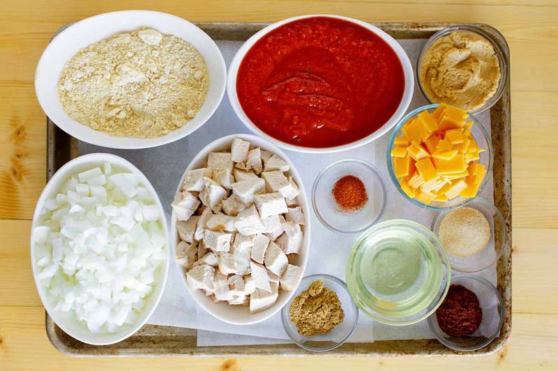 An overhead view of the ingredients for Chili's chicken enchilada soup on a baking sheet. Pictured are bowls of diced cooked chicken, diced onion, crushed tomatoes, masa harina, cubed American cheese, chicken base, oil, and various spices.