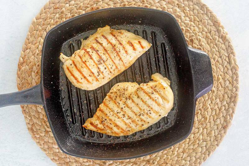 A top-down view of two seasoned chicken breasts cooking in a black grill pan, showing distinct, golden-brown grill marks on the chicken.