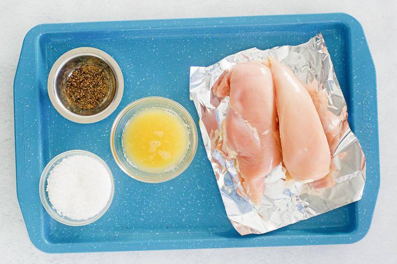 An overhead view of the simple ingredients for Chili's margarita grilled chicken on a blue tray. Pictured are two raw chicken breasts and small bowls containing margarita mix, salt, and pepper.