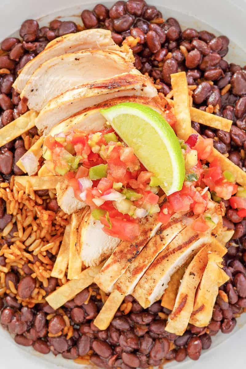 An overhead, flat-lay view of a bowl of Chili's margarita grilled chicken. The sliced chicken is served over Mexican rice and black beans and topped with pico de gallo, tortilla strips, and a lime wedge.