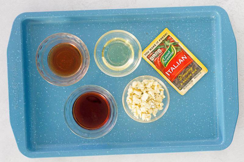 An overhead view of the ingredients for Outback blue cheese vinaigrette on a blue tray. Pictured are a packet of Good Seasons Italian dressing mix and small glass bowls containing balsamic vinegar, red wine vinegar, vegetable oil, and blue cheese crumbles.