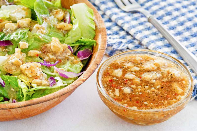 A small glass bowl of homemade Outback blue cheese vinaigrette sits next to a large wooden bowl of fresh green salad. The vinaigrette is a dark, tangy dressing with visible crumbles of blue cheese and herbs.