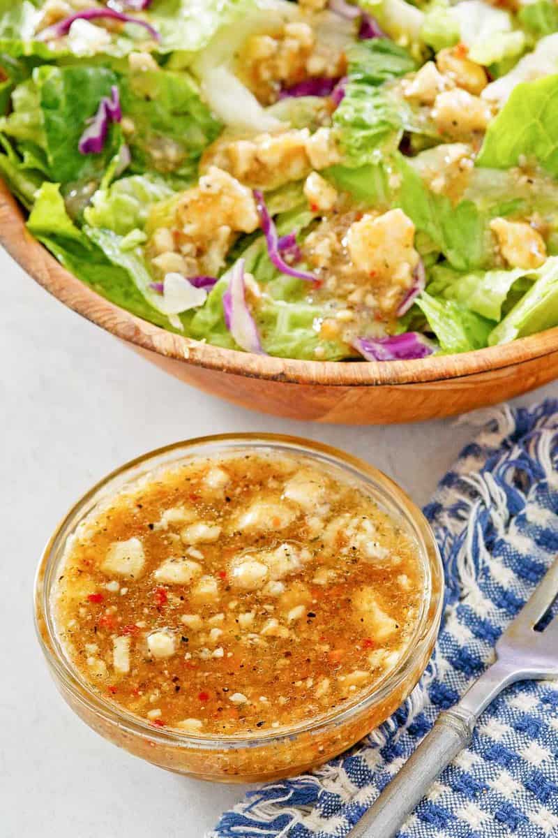 A small glass bowl of homemade Outback blue cheese vinaigrette sits next to a large wooden bowl of fresh green salad. The vinaigrette is a dark, tangy dressing with visible crumbles of blue cheese and herbs.